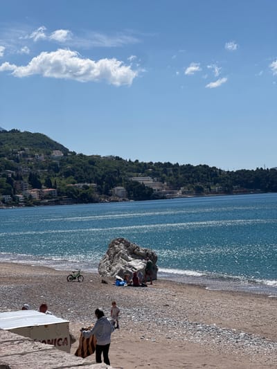 This image captures a vibrant coastal scene in Zankovići, Montenegro, under a clear, bright sky. The original image and its crops are oriented sideways, but assuming an upright view:
The upper portion of the frame features a bright blue sky dotted with scattered white cumulus clouds, indicating fair weather. Below, the sparkling blue waters of the Adriatic Sea stretch across the middle, reflecting sunlight with numerous glittering points. Gentle waves can be seen lapping onto the shore.
To the left, a steep, green hillside covered in dense trees rises from the coastline. Numerous buildings, likely homes or hotels, are nestled amongst the foliage, characteristic of a Mediterranean coastal settlement.
The foreground reveals a beach composed of both sand and pebbles. On the beach, a large, light-colored rock stands prominently near the water's edge. Several people are gathered around and behind this rock, appearing to relax. Further to the right, a person in a light blue shirt and dark pants stands beside a striped blanket on the sand. A green bicycle is parked upright near the water. In the bottom right corner, a white boat is partially visible, with the word "ANICA" clearly painted in red letters on its side. Portions of other individuals can be seen near the boat.
The scene depicts a sunny and peaceful daytime setting, showcasing the natural beauty of the Montenegrin coast combined with human leisure activities.