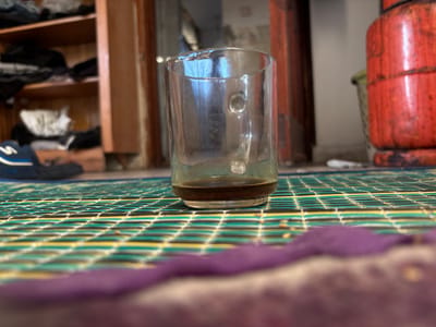In the city of Gaza, Palestinian Territories, a low-angle, eye-level shot captures a partially filled glass of dark liquid, likely coffee, resting on a woven mat. The mat features a repeating pattern of green, yellow, and black stripes. In the foreground, a blurred purple object is visible. In the background, a wooden shelf unit is filled with folded clothing, and to the right, a large red gas cylinder is present. The scene appears to be indoors, possibly in a residential setting.
