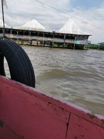 The image displays a waterside scene from the perspective of a boat on Lake Victoria, near the Dunga Beach area in Kisumu, Kenya. In the immediate foreground, the weathered red wooden gunwale of a boat is visible, partially obscuring a black rubber tire that functions as a fender. The midground features moderately choppy, brownish-green water with small waves. In the background, a large, elevated structure, identifiable as a restaurant or lodge, stands on numerous wooden stilts over the water. This building exhibits multiple prominent, sharply peaked roofs made of corrugated metal. Its lower level incorporates an open veranda with railings painted in distinct, alternating segments of red, green, yellow, and blue. Sparse green vegetation is visible at the water's edge beneath the structure. A smaller building with a green roof is partially visible to the far right in the background. The sky above is partly cloudy with patches of blue. No individuals, explicit actions, or interactions are depicted.