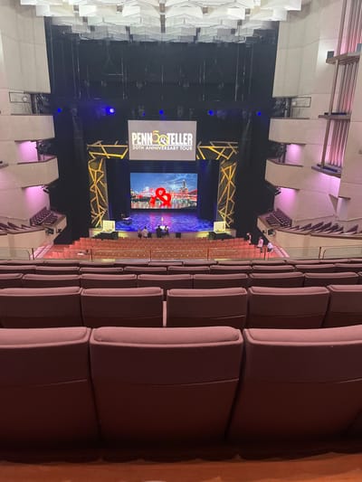 This image captures the interior of a large auditorium or theater, likely located in South Brisbane, Australia, from an elevated perspective. The central focus is a stage meticulously set for a performance.

On the stage, two prominent screens are visible. The left screen displays "PENN&TELLER 50TH ANNIVERSARY TOUR" in white and yellow text on a dark background, indicating the specific event. The right screen shows a stylized cityscape, possibly a skyline, with a large, vibrant red ampersand symbol (&) positioned in the foreground. Stage lighting, including blue spotlights, is active, illuminating the performance area, flanked by decorative gold and black geometric stage props.

Rows of empty, dark-colored upholstered seats fill the foreground and midground, sloping down towards the stage, suggesting the photo was taken before or after a show, or during a rehearsal. The seating arrangement indicates a substantial capacity for the venue. Architectural elements of the hall, characterized by angular, light-toned surfaces, are visible on the left and upper portions of the image, subtly highlighted with ambient purple lighting. The overall setting is modern and professional, designed for live entertainment.