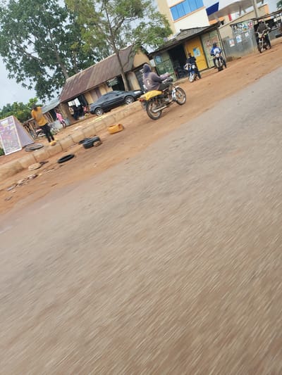 The image captures a bustling street scene in what appears to be a semi-urban or rural setting, oriented on its side. A wide, light brown, unpaved road dominates the right side of the frame, showing signs of dirt and light wear. Along the left side of the road are several simple, single-story structures, likely shops or homes. These buildings feature corrugated metal roofs and walls, with one structure displaying a distinct blue and white facade. Lush green trees flank the buildings on the far left, extending upwards towards an overcast or hazy sky.

Multiple subjects and objects populate the scene. Several motorcycles are visible; a prominent yellow motorcycle stands in the mid-ground, while another blue motorcycle is parked near one of the buildings. A black car is partially parked under a corrugated metal overhang. Several people are present, though their features are indistinct. One individual in dark grey and yellow attire is seen near the yellow motorcycle, and others are scattered near the buildings and along the roadside. Various items are strewn along the dirt shoulder, including what appears to be a black tire, a yellow jerry can, and other debris or goods. Posters are visible on some building walls, and a wooden stand with an illegible colorful sign is in the bottom left corner.

The scene depicts everyday activity, capturing a snapshot of daily life and commerce along the roadside. The time of day appears to be daytime, under an overcast sky. The overall atmosphere suggests a typical, calm day in the region, consistent with the provided location context of Vom, Nigeria.
