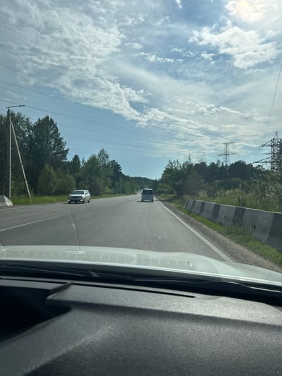 The image depicts a view from inside a vehicle traveling on a highway near Angarsk, Russia. The road is asphalt, with two lanes visible in the direction of travel and a concrete barrier separating the lanes from the opposite direction. Trees and vegetation line the sides of the road, and tall power lines and transmission towers are visible in the distance. The sky is partly cloudy, with the sun appearing to be high in the sky. Two vehicles are visible on the road ahead, one silver sedan in the left lane and a white van in the right lane. The interior of the vehicle, including the dashboard and windshield, is partially visible in the foreground.