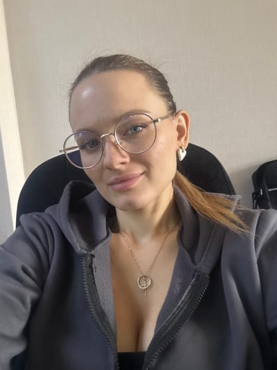 A woman is taking a selfie in a dimly lit room. She is wearing a grey hoodie, glasses, and a necklace with a cross pendant. Her hair is pulled back into a ponytail, and she is wearing heart-shaped earrings. The background is a plain, light-colored wall. There are no discernible landmarks or context to suggest the location is Irkutsk, Russia.