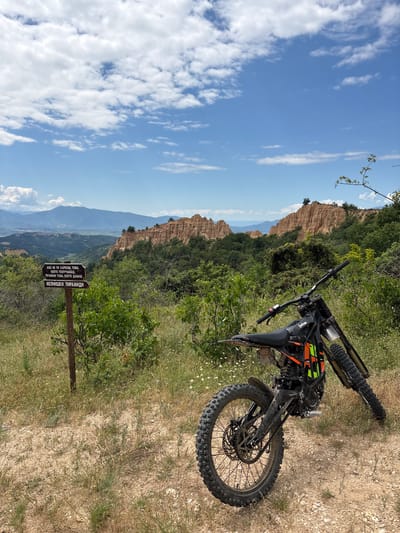 In the city of Rozhen, Bulgaria, a black dirt bike with orange and green accents is parked on a dirt path. The bike faces away from the viewer, with its rear wheel towards the foreground. Behind the bike, a wooden signpost stands on the left side of the image. The sign has two boards, the top one inscribed with text in a Cyrillic alphabet, and the bottom one with an arrow pointing to the right, indicating "MELNISHKI PIRAMIDI" (Melnik Pyramids). The background features a landscape of dramatic, eroded sandstone formations known as the Melnik Pyramids, a lush green forest, and distant mountains under a bright blue sky with scattered white clouds.
