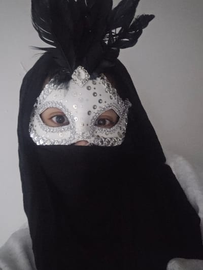 A person is wearing a white Venetian mask adorned with silver sequins and black feathers. The mask covers the upper portion of their face, revealing only the eyes. The person is draped in a black garment with a hint of a white or grey fabric underneath. The background appears to be a plain white or light-colored wall. The image is slightly tilted to the right.