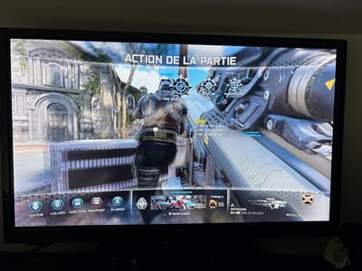 A digital photograph captures a television screen displaying a video game in progress. The game interface, primarily in French, shows "ACTION DE LA PARTIE" (Action of the Game) at the top. The scene depicts a third-person perspective of a heavily armored player character standing behind a low, light-colored concrete barrier. The character holds a large, futuristic-looking rifle over the right shoulder. To the right, a detailed view of the weapon's scope and barrel occupies a significant portion of the screen, indicating a first-person perspective or weapon focus.

On-screen display elements include score updates such as "+600" and "+100 tir dans la tête" (headshot), along with various perk icons labeled "FANTÔME," "VIGILANCE," "SANG FROID," "AVANCEMENT," and "ÉCLAIREUR." A weapon description identifies "EX-3 30M FUSIL DE PRÉCISION" (EX-3 30M Precision Rifle) at "NIVEAU 18" (Level 18) for a player at "Prestige 2, 55".

The virtual game environment features weathered stone buildings with ornate architectural details, including large arched doorways and windows, and sections overgrown with ivy. This architecture is reminiscent of historic European Gothic or Romanesque styles. This depicted environment is a fictional game map and does not represent a specific, real-world location within the city of Saint-Denis, France. The scene is static, representing a paused moment or replay within the game.