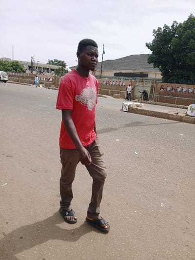The image displays a young adult Black male walking on an asphalt road in Kwekwe, Zimbabwe, under an overcast sky. He is positioned in the foreground, oriented slightly right and facing towards the camera's general direction. He wears a red short-sleeved t-shirt with a white graphic print, brown trousers, and black slip-on sandals. A silver-colored cross pendant hangs from a chain around his neck. His left arm is bent at the elbow, extended forward, and his shadow is cast to his left on the road.
In the background, to the left, a white van is partially visible, and another individual in a light blue shirt walks further down the road near a light-colored multi-story building. To the right, a low concrete wall, adorned with numerous white rectangular posters (possibly political campaign materials), separates the road from a prominent building featuring a dark grey tiled roof and light-colored walls. A green flag on a pole stands near this building, with a large green tree behind it. Two additional individuals are visible near the building's entrance: one standing in a white shirt, and another crouched on the ground. The scene depicts an urban environment during daylight.