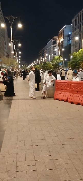 This nighttime image depicts a bustling pedestrian street in Medina, Saudi Arabia, characterized by modern multi-story buildings illuminated by numerous ornate streetlights. The ground is paved with light-colored interlocking tiles. Numerous individuals are present on the thoroughfare. Many men are observed wearing traditional white thobes, some with head coverings, while many women are dressed in black abayas and head coverings. In the left foreground, a man in a white thobe stands facing slightly right. To his left, a woman in a black abaya kneels or stoops, interacting with an unseen subject. In the right mid-ground, a man in a white thobe is seated on a light-colored cylindrical stone bollard, his right arm resting on his knee, positioned next to two stacked orange-red plastic construction barriers. Other people are seen walking in the background in various directions.
