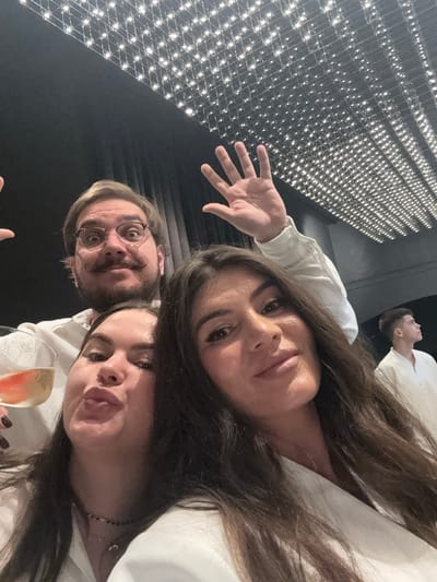 The image shows a selfie taken by three individuals, two women and one man, who are dressed in white clothing. The man is in the background, wearing glasses and a distinctive mustache, with his hands raised as if waving. The two women are in the foreground, with one holding a glass of champagne. The background appears to be an indoor venue with a dark ceiling illuminated by a grid of small lights. A blurry image of another person in white clothing can be seen in the far right background. The scene does not provide any specific details to suggest a location in Irkutsk, Russia.