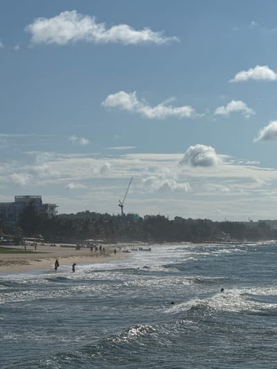 The image captures a coastal scene in Phan Thiết, Vietnam, with a view of the ocean, beach, and a town in the background. The foreground is dominated by the ocean, with choppy waves rolling towards the shore. Several people are visible in the water, some appearing to be swimming or wading. Further back, the sandy beach is populated with individuals walking and relaxing. Beach umbrellas and some watercraft are scattered along the shoreline. In the middle ground, a dense line of palm trees separates the beach from the town. Buildings, including a large multi-story structure and a construction crane in the distance, are visible behind the trees, suggesting ongoing development or infrastructure projects. The sky above is a clear blue with scattered white clouds, indicating a bright, possibly sunny day.