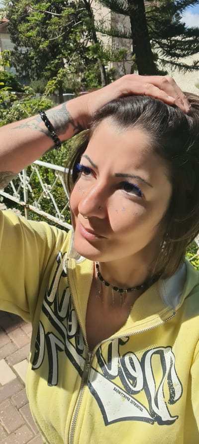 A woman with blue eyelashes and facial glitter poses outdoors with her hand on her head. She wears a yellow zippered hoodie with black graphic text. She also has a black beaded necklace and bracelet. In the background, there's greenery, a white railing, and a glimpse of trees. The image appears to be taken in Nahariyya, Israel, as part of the image was used by this city.