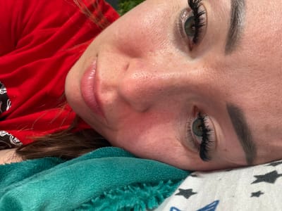 The media file is a close-up, vertically oriented photograph of a person's face, primarily focusing on their eyes, nose, and lips. The subject appears to be lying down, with their head tilted upwards. They have noticeable false eyelashes, light-colored eyebrows, and freckles across their nose and cheeks. Their skin shows some redness, particularly on the cheeks. The subject's eyes are green, and they are looking slightly upwards and to the left. The lips are painted with a glossy pink lipstick.
Behind the subject's head, there are soft, textured fabrics. One is a white fabric with a pattern of dark gray stars, and another is a fluffy, teal-colored material. The subject is wearing a red garment, possibly a shirt, which is draped around their body and fills a significant portion of the lower right side of the frame. A small section of dark hair is visible.
The lighting suggests it is daytime, possibly outdoors given the dappled shadows and the general brightness. The overall impression is one of a casual, relaxed pose. There is no visible text in the image. The location context provided is Старые Туймазы, Russia, but there are no specific visual cues within the image to confirm this location.