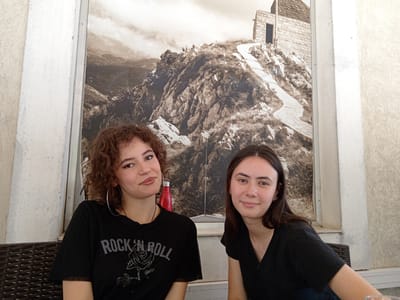 Two young women are seated at a table in an outdoor cafe. They are positioned in front of a large mural depicting a mountain landscape with a stone building at the summit and a winding path. The woman on the left has curly brown hair and is wearing a black t-shirt with a "Rock N Roll" graphic and a rose. She has large hoop earrings. The woman on the right has long, dark brown hair and is wearing a black t-shirt. The cafe appears to be located in Cetinje, Montenegro, as indicated by the mural which is a well-known landmark at Lovcen. The seating area is furnished with dark wicker chairs.