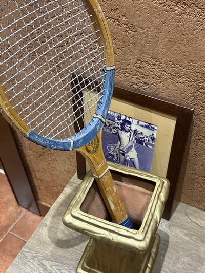 This image depicts a display setup featuring a vintage tennis racket and a framed photograph.
In the foreground, a wooden tennis racket is positioned vertically within a square, gold-painted, ornate stand or vase. The racket head exhibits a worn blue painted section around its rim. Its strings are white with faint blue interweaving, forming a tight grid. The racket's throat features a faded gold emblem, and the wooden handle shows a blue and red striped grip near its base where it enters the stand. The stand's interior is a dull reddish-brown.
Behind the stand, a dark brown wooden-framed photograph rests against a textured wall. The photograph is a black-and-white or sepia-toned image with a blue cast, depicting a male tennis player on a court, racket in hand, with spectators visible on bleachers in the background.
The display rests on a surface of light-colored wooden planks. To the far left, terracotta-colored square floor tiles are partially visible. The wall behind the display is uniformly textured in an earthy brown or terracotta color, resembling stucco.
The specific address or interior space within Cukarica Urban Municipality, Serbia, is not ascertainable from the visual information provided.