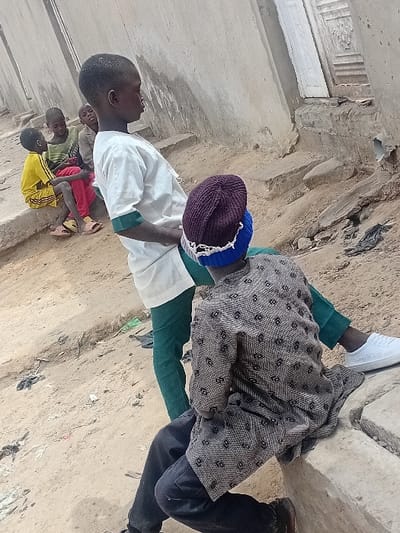 The scene depicts five young males congregating outdoors in the city of Ayama, Nigeria. In the foreground, an individual sits on a concrete ledge with their back to the viewer, wearing a dark purple and blue knitted beanie, a grey patterned long-sleeved shirt, and dark trousers. Their right foot, clad in a white sneaker, rests on the ledge. Adjacent to them, another male stands, facing right, dressed in a white long-sleeved top with green cuffs and green trousers. Further in the background to the left, three other males are seated on a lower concrete ledge. One wears a yellow top and shorts with pink sandals, another a green and black striped long-sleeved top, and the third a light brown long-sleeved top. The ground is unpaved dirt with scattered debris, including dark plastic. Unpainted concrete building walls form the background, with a decorative light-colored wooden door visible on the right. The overall environment appears to be an urban or semi-urban outdoor area.