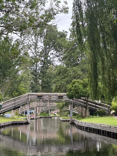 The image depicts a serene canal scene in Giethoorn, Netherlands, a village renowned for its waterways and lack of roads. A wooden bridge, with its distinctive ramped approach, spans the dark, reflective water. The bridge is adorned with flower baskets, and on its railing, a pair of flip-flops are casually placed. On the left bank, a blue boat and a red canoe are visible near the grassy edge, suggesting a peaceful, recreational environment. Lush green trees, including a weeping willow on the right, frame the scene, their foliage creating a dappled canopy overhead. The water's surface mirrors the bridge and the surrounding greenery, creating a tranquil, almost painterly effect. The overcast sky lends a soft, diffused light to the scene, contributing to the overall sense of calm.
