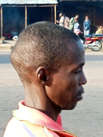 A medium shot captured from the side shows the upper body and head of a Black man. The man is facing right with his eyes closed, and appears to be in motion. He has short, dark hair and is wearing a pink shirt with a yellow collar. In the background, several people are gathered near a building with a corrugated metal roof. A red and white motorcycle is parked to the right of the group. The scene appears to be outdoors, possibly on a street or in a marketplace. The overall lighting suggests it is daytime.