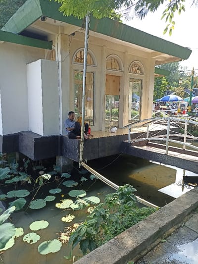 This outdoor scene, set in Mataram, Indonesia, features a light-colored building with a distinctive green roof overhang, situated alongside a body of water. The building stands on an elevated, dark-colored foundation, and its facade is characterized by multiple tall, arched windows, some of which appear to be boarded up or covered from the inside with a light-colored material.
Two people are visible on the building's elevated platform. A man, dressed in a blue shirt and dark pants, is seated and leaning against the building's wall. Beside him, a person, possibly a child, is also sitting, partially obscured, wearing red clothing. They appear to be resting or relaxing in the bright daylight. A small white object is on the platform near them.
The water in the foreground, and extending beneath the building and a bridge, is dark and adorned with numerous large, round green lotus leaves. A white, arched pedestrian bridge with railings spans a section of the water, connecting to the building's platform. Beyond the bridge, in the background, several colorful market umbrellas (blue, red, and white) and what seem to be market stalls or vendor setups indicate a lively public or commercial area. Various green plants and trees contribute to the natural setting.
The image was captured during the daytime, under clear, sunny weather, as evidenced by the bright illumination and distinct shadows. The overall atmosphere suggests a public recreational space or park adjacent to a marketplace. No discernible text is visible within the image.