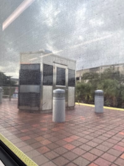 A hazy view from a train window reveals an exterior scene. A modern building with large windows sits beside a platform paved with red and orange brickwork. Two cylindrical gray trash receptacles are placed near the building. Overcast skies and muted greenery are visible in the background, partially obscured by the window's imperfections. The scene is slightly rotated.
