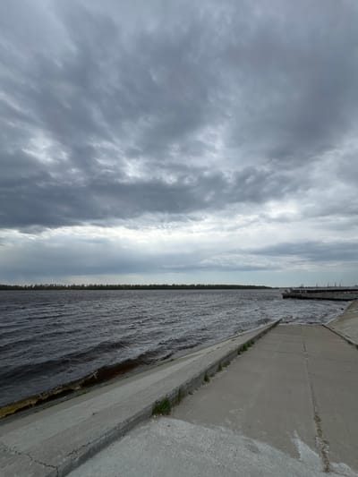 The image shows a concrete embankment or dam on a cloudy day. The water is dark and choppy. Some greenery grows along the edge of the concrete structure. The sky is filled with heavy, gray clouds. The photo was taken in Nizhnevartovsk, Russia.