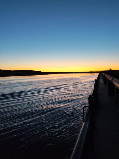 A wide shot captures a serene scene at dusk from the perspective of a boat's railing. The sky transitions from a deep blue at the top to a vibrant orange-yellow near the horizon, where a silhouetted treeline marks the distant shore. Gentle ripples disturb the dark water, reflecting the warm hues of the sunset. The boat's metal railing runs along the right side of the frame, leading the eye towards a distant structure on the far shore. The overall atmosphere is peaceful and still.