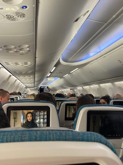 The scene depicts the interior of a commercial aircraft cabin, viewed from a passenger's perspective looking forward down the aisle. The environment is consistent with an airplane during flight, potentially associated with air travel to or from Seeb Province, Oman, where Muscat International Airport is situated.

Multiple rows of passengers are seated, primarily visible from behind, occupying economy-class seats upholstered in turquoise and white fabric. The cabin ceiling features white panels with individual air vents, reading lights, and passenger call buttons. Blue ambient lighting illuminates the upper portion of the cabin. An "EXIT" sign is visible further down the aisle, indicating emergency egress.

In the foreground, the seatback directly in front of the viewer contains an active in-flight entertainment screen. This screen displays an image of a young woman, shown from the chest up, wearing a dark head covering (hijab) and facing directly forward. The background within the screen's display appears to be an indoor setting with architectural elements. Another seatback screen, displaying indistinct content, is visible to the right. No active interactions between passengers are discernable.
