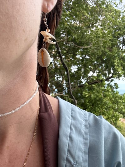This close-up media file features a person's shoulder and neck area, adorned with jewelry. The subjects are a person and their accessories: a shell earring with a white cowrie shell and a delicate pearl necklace. The setting appears to be outdoors, with lush green trees and dappled sunlight suggesting a natural environment. The lighting indicates it is daytime, possibly late morning or early afternoon, given the bright illumination and the subtle shadows.
The person's skin is visible, with some freckles and a smooth texture. The jewelry is the focal point of the image: the shell earring dangles from the ear, showcasing intricate detailing on the small shell charm, and the pearl necklace rests gently on the collarbone. A hint of blue fabric, possibly a shirt or blouse, is visible on the upper right side of the frame, adding a touch of color and contrast to the otherwise natural tones of the scene. The background is softly blurred, emphasizing the foreground details. There are no visible text elements in this media file. The overall impression is one of casual elegance and a connection with nature, possibly during a relaxed outing in a tropical or subtropical location like Baan Thurian, Thailand.