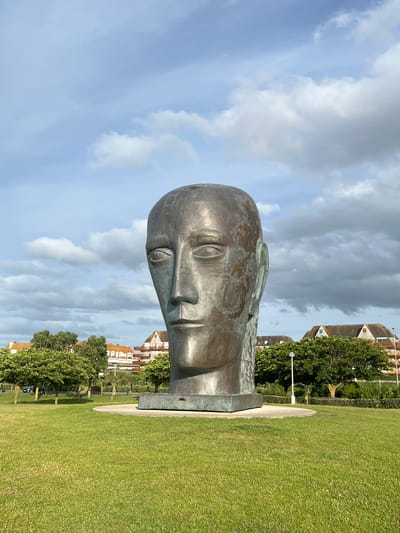 This is a frontal, eye-level, outdoor, medium-shot photograph of a large, bronze sculpture of a human head. The sculpture is positioned centrally in the frame and is the primary subject. The head is depicted in a neutral, stoic expression, with closed eyes and an unexpressive mouth. The bronze material shows signs of weathering, with green patina developing in some areas. The sculpture rests on a square, grey concrete base, which is on a circular concrete platform.
Behind the sculpture, the environment consists of a well-maintained, bright green lawn. In the middle ground, there are several trees with lush green foliage, and beyond them, buildings with orange-tiled roofs are visible. The sky above is a bright blue, dotted with scattered white and grey clouds. A street lamp is visible to the right of the sculpture.
This sculpture is known as "La Tête Carrée" or "The Square Head" and is located in the city of Deauville, France, specifically in the Place du Dauphine, which is a public square near the waterfront.