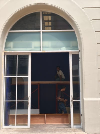 Two men are working on a construction project inside a large arched doorway in South Jakarta, Indonesia. The doorway has a white frame and features a large arched window above a set of glass double doors. The upper part of the doorway is filled with frosted glass panes, while the lower part consists of clear glass doors with a white grid.
One man, wearing a light-colored t-shirt and blue jeans, is crouched on a wooden platform inside the doorway, looking to his left. He appears to be supervising or inspecting the work. The second man, wearing a dark t-shirt and blue jeans, is standing further inside the doorway and to the right. He is holding a mobile phone in his right hand and looking down, possibly taking a break or communicating with someone.
Behind the men, scaffolding structures are visible, along with dark blue tarpaulin or fabric covering the back wall of the interior space. Wooden shelves or boxes are arranged on the floor within the doorway. The exterior of the building is a light beige color, with architectural molding around the arched doorway. The ground in front of the doorway is concrete.