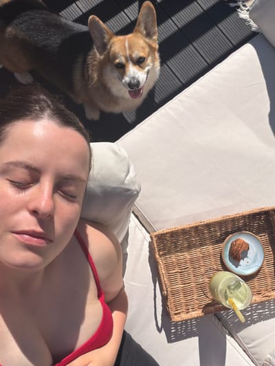 A fair-skinned woman wearing a red top lies on a beige cushioned lounger. A corgi dog with its tongue sticking out sits to her left. A woven tray containing a blue plate with a pastry and a glass of a green beverage with a straw rests on the lounger beside the woman. The scene appears to be outdoors, and there is a glimpse of dark wood-like flooring to the left of the lounger. There is no visible evidence to suggest the location is Gangloff, Germany.
