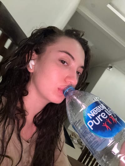 A woman with dark hair and fair skin is drinking from a "Nestle Pure Life" water bottle. She is wearing a white wireless earbud in her left ear. The picture appears to be taken at an angle, with a partial view of a room in the background, including a ceiling fixture and a wooden slatted structure. It is possible this image was taken in Dalung, Indonesia.