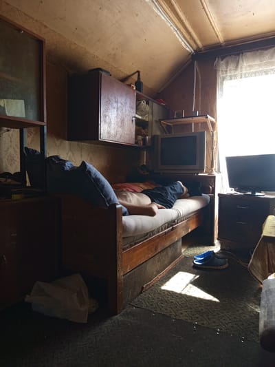 The image depicts a dimly lit, cluttered room that appears to be a bedroom or living space. A person, partially visible from the chest down, is lying on a narrow wooden bed, wearing dark pants and black socks. The bed is covered with a white sheet and a dark blue pillow. To the left of the bed, a wooden cabinet with a glass-fronted section is visible, containing some items. Above the bed, a wooden cabinet is mounted on the wall. In the background, a window with white lace curtains is letting in bright sunlight, casting shadows across the floor. Next to the window, a modern television is placed on a dark wooden dresser. Another older television set is positioned on a shelf above the bed. The floor is covered with a dark, textured rug, and a pair of blue clogs are placed near the foot of the bed. A crumpled white plastic bag is visible under the bed. The room's walls and ceiling are paneled with wood, and the overall impression is one of a lived-in, somewhat disorganized space. The scene is located in Valmiera, Latvia, within a residential building.