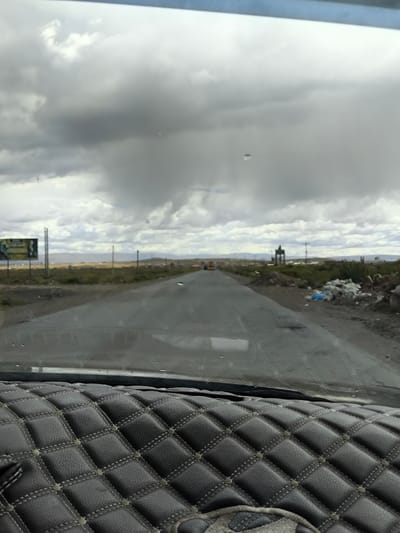 From a car's perspective, a gray, rain-spattered road stretches ahead beneath a heavy, overcast sky. Distant hills fade into the horizon under the gloomy light. Patches of vegetation line the roadside, with litter visible on the right. A distant vehicle is seen on the road, which appears wet. The car's dashboard, covered in quilted material, fills the foreground.