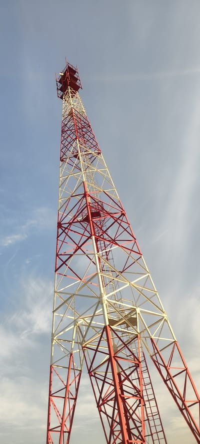 A tall, red and white lattice tower, likely a telecommunications tower, stands against a light blue sky with wispy clouds. The tower is viewed from a low angle, emphasizing its height and structure. The lower portion of the tower is red, transitioning to white with red accents higher up, and finally culminating in a red top section with some equipment visible. A ladder runs along the side of the tower for access. The scene appears to be an open area, consistent with an airport environment, possibly at Yemelyanovo Airport near Yemelyanovo, Russia. No people or specific actions are depicted.
