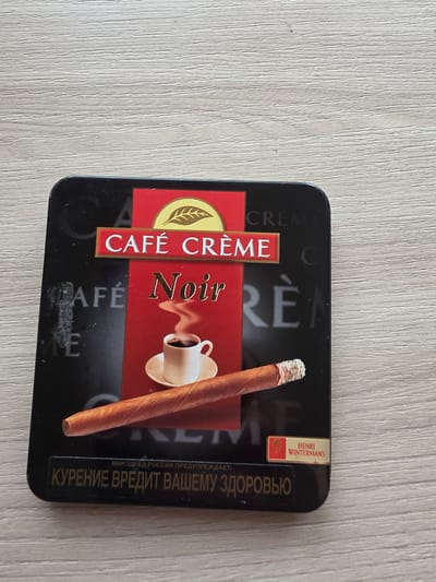 The image shows a dark, rectangular tin with a lid. The lid is decorated with text and images related to coffee and cigars. Prominently displayed are the words "CAFÉ CRÈME" in white letters on a red background, and "Noir" in a stylized gold font. Below this, there is an illustration of a steaming coffee cup and a cigar. In the bottom section of the lid, there is text in Russian that translates to "Smoking is harmful to your health." A small red and gold label in the bottom right corner reads "HENRI WINTERMANS." The tin is placed on a light-colored, wooden surface with visible grain. The scene is static and indoors. No specific location in Parshino, Russia, can be identified from this image.
