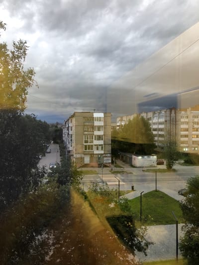 This is a view from a window in Solikamsk, Russia, overlooking a street with apartment buildings, trees, cars, and a cloudy sky. In the foreground, reflections of trees and sunlight are visible on the windowpane. The street runs between two multi-story apartment buildings, one on the left and one on the right. Several cars are parked along the street. There is a pedestrian crossing on the street. Green grass areas and streetlights are present on the sidewalk. The sky is overcast with dark clouds.