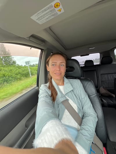 A woman is shown from the chest up, sitting in the passenger seat of a car, looking at the camera. She has light skin, blue eyes, and brown hair pulled back in a ponytail. She is wearing a light blue jacket over a white sweater. A gray seatbelt is fastened across her chest. The car's interior is visible, including black leather seats and a sunroof overhead. Outside the car window, green trees and vegetation can be seen, suggesting a rural or suburban setting. The sky appears overcast. There is no discernible indication of the city of Kopeysk, Russia.