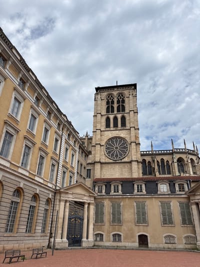 This image shows the exterior of a historic building in Lyon, France, likely the Primatiale Saint-Jean-Baptiste Cathedral, given the architectural style and the inscription "PRIMATIALE" above an ornate gate. The scene is captured from a slightly low angle, looking up towards the imposing stone structure.
On the left, a building with a yellow facade and white decorative trim stretches across the frame. It features multiple rows of tall, arched windows with grey panes. The facade is constructed from stone blocks.
To the right of this building, a tall, stone tower dominates the center of the image. This tower has Gothic architectural elements, including pointed arch windows and decorative stonework. A large, circular stained-glass window, a rose window, is visible on the tower's facade. The tower is connected to a larger, longer stone building with arched windows and spires, characteristic of a cathedral.
In the foreground, there is a reddish-brown ground surface, possibly a courtyard or plaza. Two simple wooden benches are placed on this surface in the lower left corner of the image. Between the yellow building and the cathedral, there is an entrance with large, dark, ornate metal gates framed by stone pillars and topped by a pediment with the "PRIMATIALE" inscription.
The sky above is overcast with a mix of grey and white clouds, suggesting a diffused daylight. No people are visible in the image. The overall impression is one of grandeur and historical significance.
