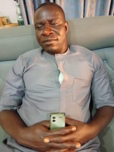 A single adult male is depicted seated indoors in what appears to be a residential or lounge setting within the city of Jimeta, Nigeria. The individual, with a dark complexion and short hair, is positioned centrally in the frame, directly facing the camera. He wears a light grey or lavender-colored long-sleeved tunic featuring a high collar, a partial button placket, and a left chest pocket. His eyes are partially closed, suggesting a squint or relaxation, and his mouth is closed. Both hands are clasped together in front of him, resting at waist level, and holding a light gold-colored Samsung flip phone. The phone is shown from its rear, displaying two circular camera lenses and the brand logo. He is seated on a light grey upholstered sofa, which features visible tufting on its backrest to the right. The background includes a textured or subtly patterned curtain behind his left shoulder and a decorative white object, possibly a lamp or vase, on a surface further to his left.