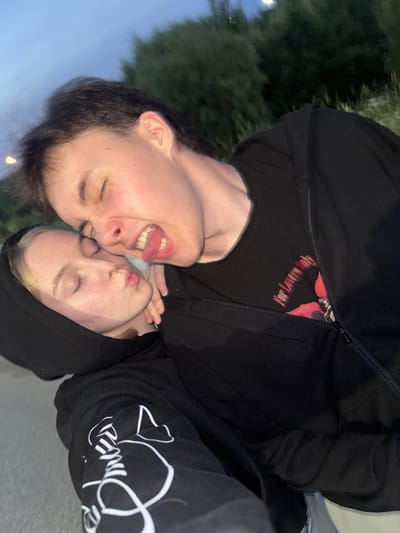 The image shows a close-up, portrait-style selfie of two individuals. The person in the foreground has their eyes closed and their tongue sticking out playfully. The second person is positioned behind and slightly to the side of the first, also with their eyes closed and their lips pursed, as if about to kiss. Both individuals are wearing black hoodies. The first person's hoodie has a red floral graphic and text that reads "For Lovers Only" printed on the chest, visible through the partially unzipped outer layer. The second person's hoodie has a white graphic design on the sleeve. The setting appears to be outdoors, with blurred greenery in the background and a pale sky. The lighting suggests it could be late afternoon or early evening. The overall mood is intimate and playful. The location context provided is Ryabinino, Russia.