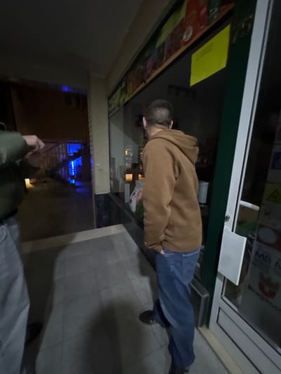 The image captures a nighttime outdoor scene in Bragança, Portugal, on a paved surface outside a commercial establishment.
Two individuals are present. On the right, a person is seen from behind, facing a shop window. This individual wears a brown hooded sweatshirt and blue jeans, with their right hand inserted into a pocket. Their left hand holds a white object or package near the lower edge of the shop window.
The storefront features a large glass display window and an adjacent glass-paneled door with a white frame. Above the display window, a colorful banner depicts various products, and a yellow rectangular sign is affixed to the upper right corner of the window. The address "Nº 15" is visible above the window. A white rectangular sign, displaying "MB" and "Bragança" within a red border, is attached to the glass door on the right.
To the left, a second person is partially visible, showing their upper torso, left arm, and left leg. This individual wears a dark olive-green jacket or hoodie and light grey pants. Their left hand is raised in front of their chest.
In the background, behind the left individual, an outdoor staircase leads to a building. A prominent blue light source illuminates the area behind and beneath this staircase, creating reflections on the ground. The ground is a light grey tiled surface, exhibiting shadows cast by the figures and ambient structures.