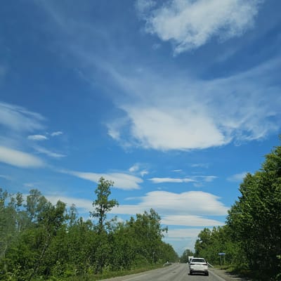 The media file shows a view from a car driving along a road lined with lush green trees on either side. The road itself appears to be asphalt, and there is a white vehicle in front of the camera. The sky is a bright blue with wispy white clouds, suggesting a sunny day.
The perspective is from the driver's seat or passenger seat of a vehicle, indicating movement along the road. The tall, dense trees suggest a rural or natural setting, likely a forest or a heavily wooded area. The presence of a road and a vehicle points to travel or a journey.
A blue sign with white Cyrillic text is visible by the side of the road. While the text is partially obscured and difficult to read clearly, the location context of Petropavlovsk-Kamchatsky, Russia, suggests it is likely a road sign indicating a place name or direction.
The overall mood is peaceful and serene, with the bright sky and green foliage evoking a pleasant outdoor experience. The time of day appears to be daytime, given the bright sunlight and clear sky. The weather seems to be fair and warm, suitable for driving and enjoying the scenery.