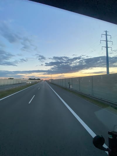 The image is a vertical, eye-level shot of a highway in Węgrzce Wielkie, Poland, taken from inside a vehicle, likely at dusk. The foreground shows the asphalt road with white lane markings. In the middle ground, vehicles are visible on the highway, and on both sides of the road are tall, gray sound barriers. Beyond the sound barriers, a power line pylon is visible on the right, and trees and buildings can be seen in the distance on the left. The sky is a mix of blue and gray clouds, with a warm orange glow near the horizon, suggesting a sunset. The overall impression is a serene, late-day drive on a highway.