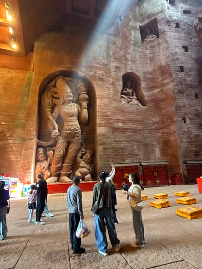 The image depicts a cavernous interior space, possibly a religious site or historical monument, likely located in Shizhong, China. The walls are rough and textured, exhibiting carved reliefs of figures, including a large central carving of a reclining figure, presumably a deity or important historical personage. Several people are present, observing the carvings. They stand on a paved floor in front of the carvings, suggesting they are visitors or pilgrims. The lighting is somewhat dim, with a bright beam of light illuminating a portion of the wall. Other elements include ornate, rectangular decorations lining a wall.