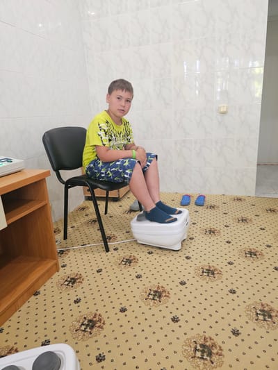 A young boy sits on a black chair in a room with tiled walls and a patterned carpet. He is wearing a bright yellow t-shirt with a graphic design and blue shorts with a white pattern. His feet are placed on a white foot massager. Blue slip-on shoes are positioned on the carpet next to the massager. To the left of the boy, a wooden shelf unit holds a medical device. The room appears to be a clinic or spa, possibly located in Lermontovo, Russia, given the visual cues which are consistent with common interior designs found in such facilities in the region.
