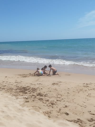 The image shows three children playing on a sandy beach near the ocean. The children are in the middle ground of the image, with the ocean and sky in the background. One child is on their hands and knees, facing away from the camera, wearing a multicolored swimsuit. Another child is kneeling to the right of the first child, facing the same direction, wearing a dark swimsuit. The third child is kneeling in front of them, facing the camera, wearing patterned shorts and is shirtless. The beach is covered in sand, with some seaweed scattered around. The ocean is a vibrant blue-green, with white waves crashing on the shore. The sky is clear and blue. The scene is set in a coastal environment, typical of beaches found in Rota, Spain.