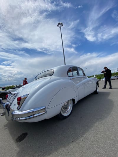 A white vintage Jaguar E-Type is parked outdoors on an asphalt surface under a cloudy sky. The car is viewed from the rear passenger side, highlighting its curved body, chrome bumper, and whitewall tires. A person in a dark jacket and pants is standing to the right of the car, holding a camera and appearing to take a photograph of the vehicle. In the background, another person in a red jacket is visible near a different car. A tall light pole with multiple lamps stands prominently in the sky. The scene suggests an outdoor car show or gathering.