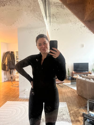 A female subject is depicted taking a mirror selfie in an interior residential setting located in Roubaix, France. The subject, an adult female with dark hair pulled back, wears a black, long-sleeved athletic top with a quarter-zip and thumbholes, and matching dark athletic leggings marked with a white "E" logo on the left hip. She holds a black smartphone in her right hand, directing its camera toward her reflection, while her left hand rests on her hip. She exhibits a subtle smile. The mirror surface displays numerous small, dark speckles and imperfections. The reflection captures a multi-room dwelling. To the reflected left, a hallway with wooden flooring contains a dark coat on a rack and a clear, metal-framed chair. A white shelving unit is visible beyond. To the reflected right, a living room area features a light-colored sofa, a dark wooden media console with a flat-screen television, and a tall cactus plant in a dark pot. A wooden staircase with a banister is present in the upper right quadrant of the reflection. A light-colored, patterned rug is visible on the floor in front of the subject.