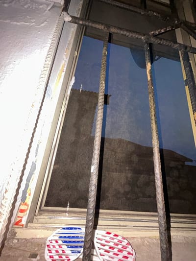 The scene depicts an interior view looking outwards through a secured window in Ilishan, Nigeria. Heavy, weathered rebar security bars with visible corrosion are prominently positioned in the foreground, installed vertically and horizontally across the window opening. Behind the bars, a dark mesh insect screen is partially visible, showing some dislodgement or damage, particularly in its upper portion. Through the screen and window glass, a dark, indistinct exterior environment is visible, possibly suggesting nighttime or a deeply shadowed area. Faint reflections with a bluish tint are present on the glass. A dark, corrugated-like structure is faintly discernible through the screen. Below the window, on an interior sill or ledge, two pairs of slatted novelty sunglasses are placed: one with red, white, and blue stripes, and the other with red and white stripes. To the immediate left of the window frame, a light-colored wall exhibits peeling paint and remnants of red-orange markings. No individuals, actions, or interactions are depicted.