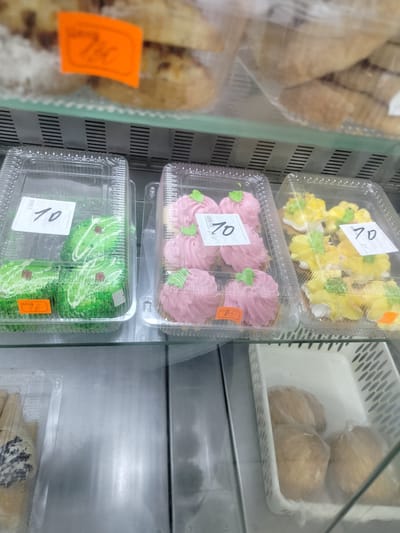 The image shows a retail display case filled with various baked goods. In the foreground, there are several clear plastic containers holding individual portions of sweets. One container on the left contains green-colored cakes with white frosting and small brown decorative elements, with a price tag of "10". To the right of this, another container holds pink frosted cakes, with a price tag of "70". Further to the right, a container holds yellow frosted cakes, also with a price tag of "70". In the background, blurry images of other baked goods, possibly bread or pastries, are visible. An orange price tag with handwritten text is also visible in the upper left background. The display case appears to be in a store. There are no people visible in the image. The location is specified as Yashkino, Russia.